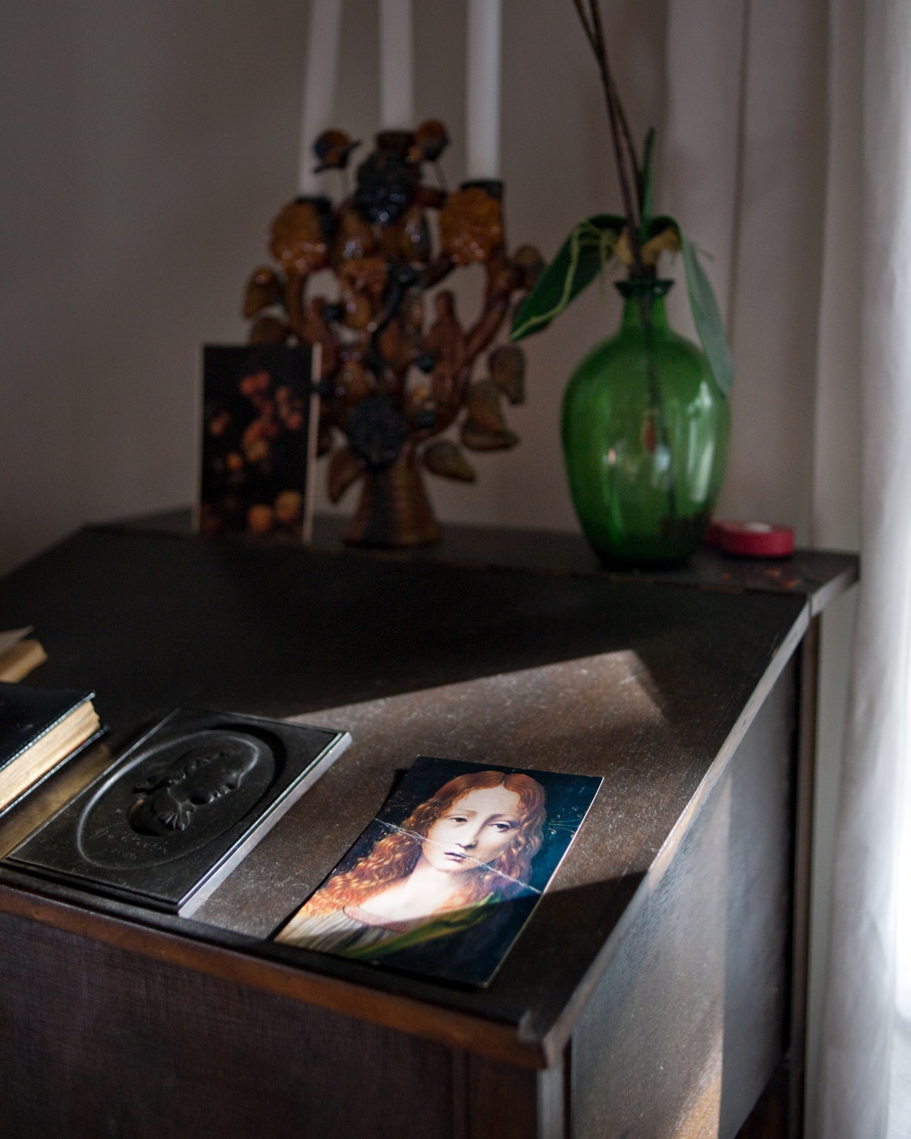 A desk displaying art books, a portrait print, decorative objects and a green glass vase with dried flowers.