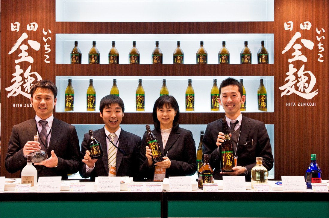 Four Japanese sales staff in business attire smiling behind a display counter with bottles of Hita Zenkoji sake and shelving.
