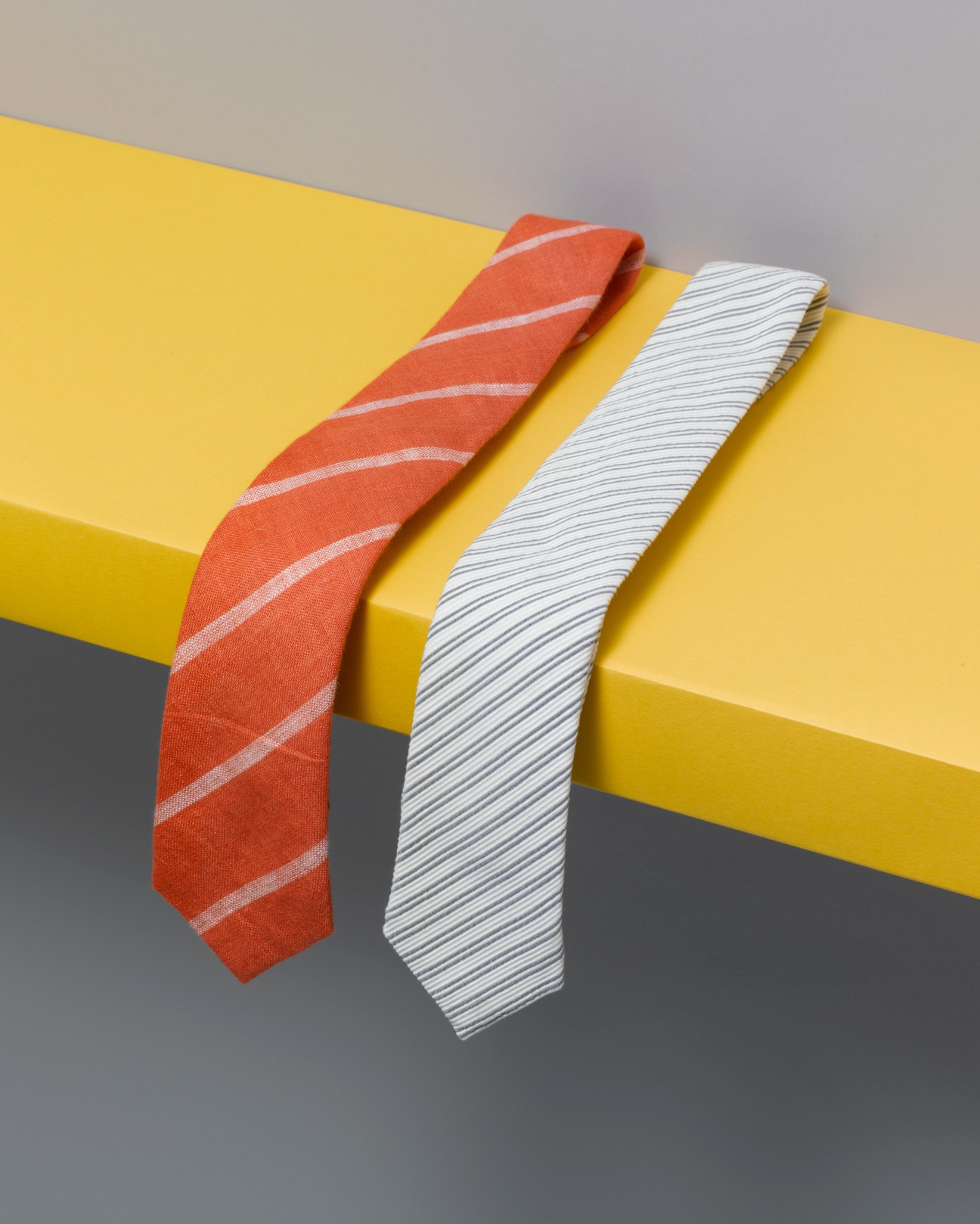 Two hand-made ties by Alexander Olch displayed on a yellow geometric form: one orange with diagonal stripes, one white with fine lines.