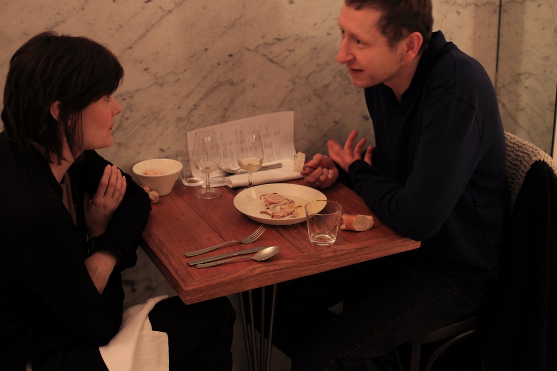 Two diners converse at a wooden table set with plated food and wine glasses at Le Dauphin restaurant.