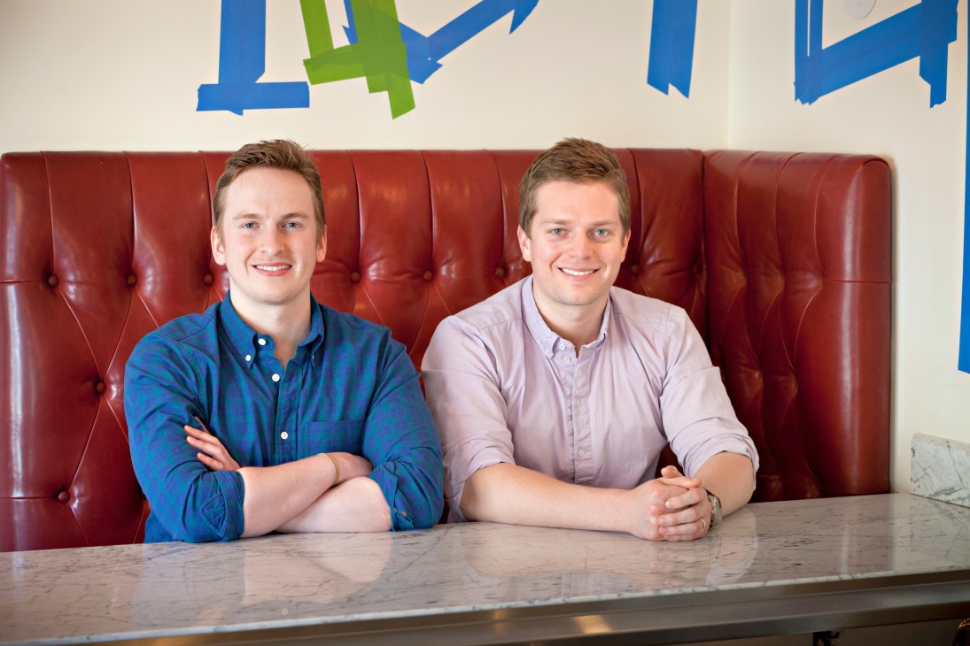 Two men seated at a counter in front of a red upholstered booth, with blue and green graphic art on the wall behind them.