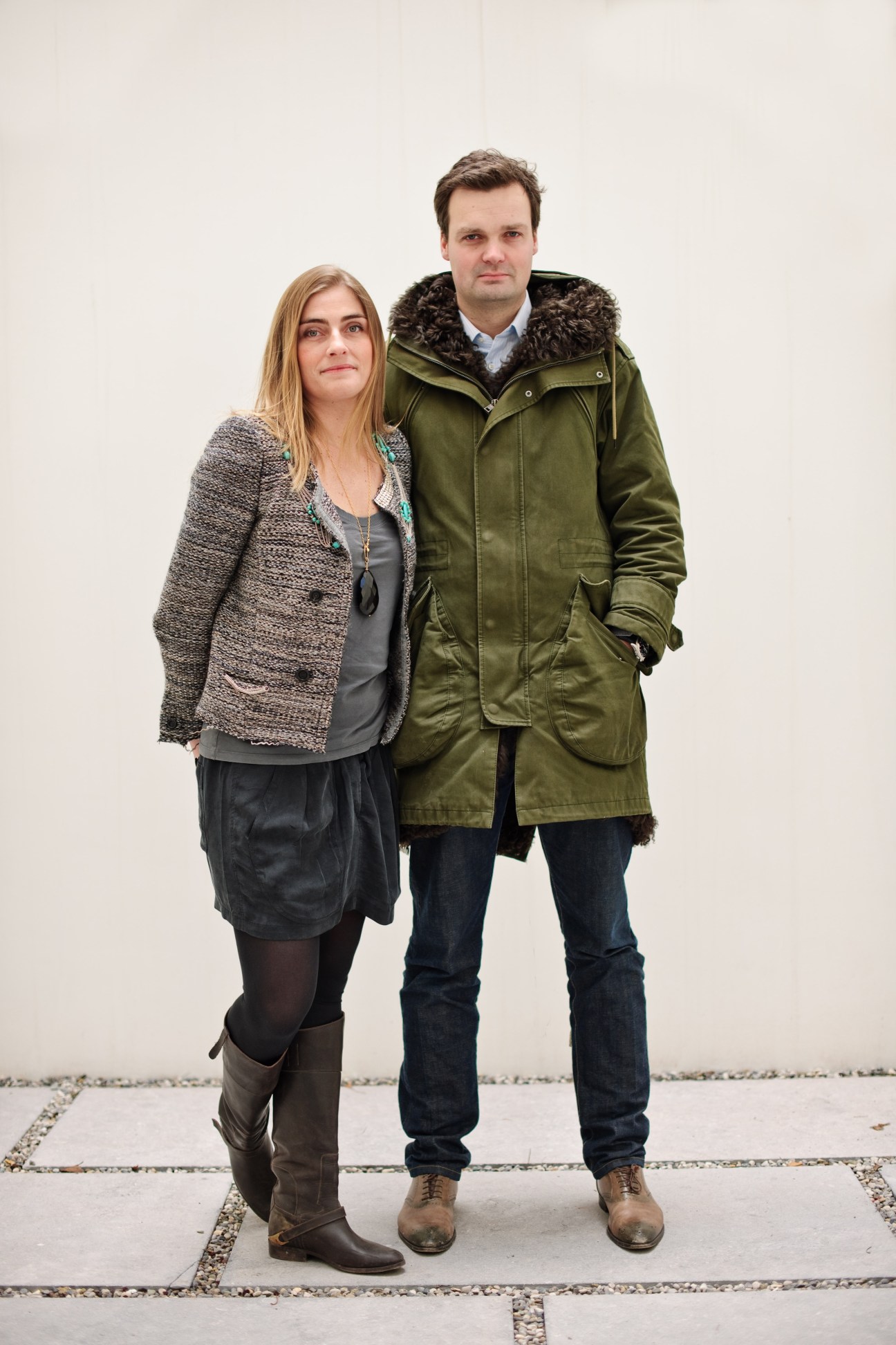 Two people standing against a white wall, woman in grey blazer and man in olive parka jacket.