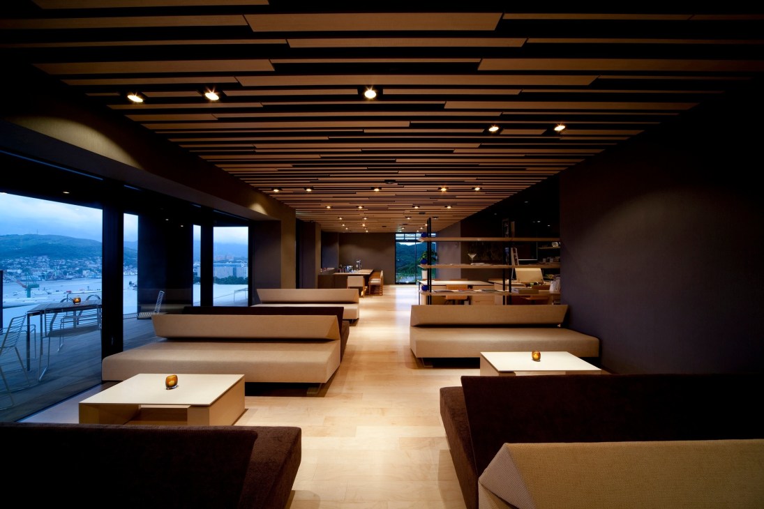 Modern hotel lounge with floor-to-ceiling windows overlooking a city at dusk, featuring minimalist furnishings and wooden ceiling panels.