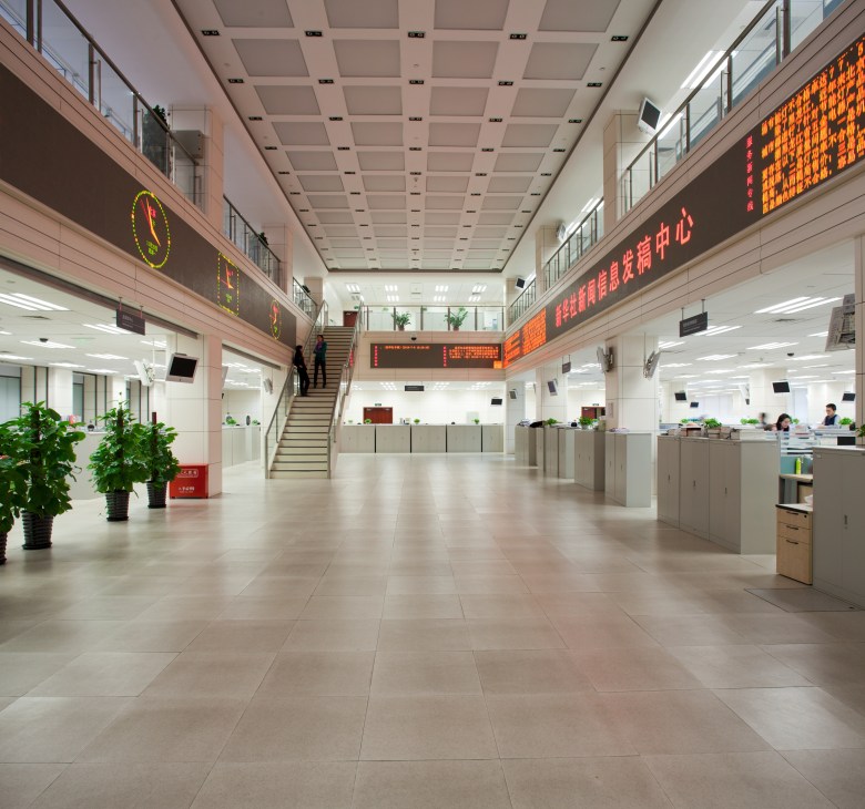 Modern newsroom interior with open floor plan, orange LED ticker displays, potted plants, and workers at desks.