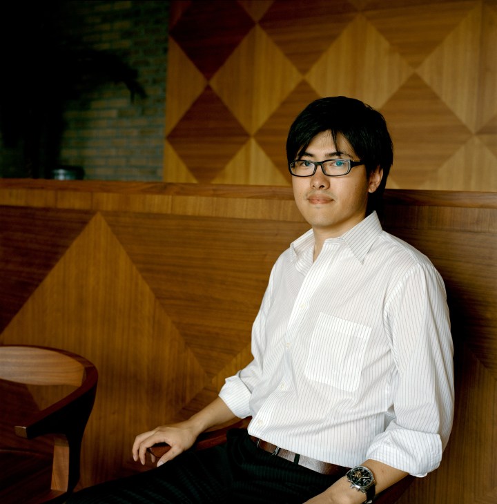 Furniture Labo chairman Yuichiro Hori sitting on a wooden bench against a geometric wood-paneled wall.
