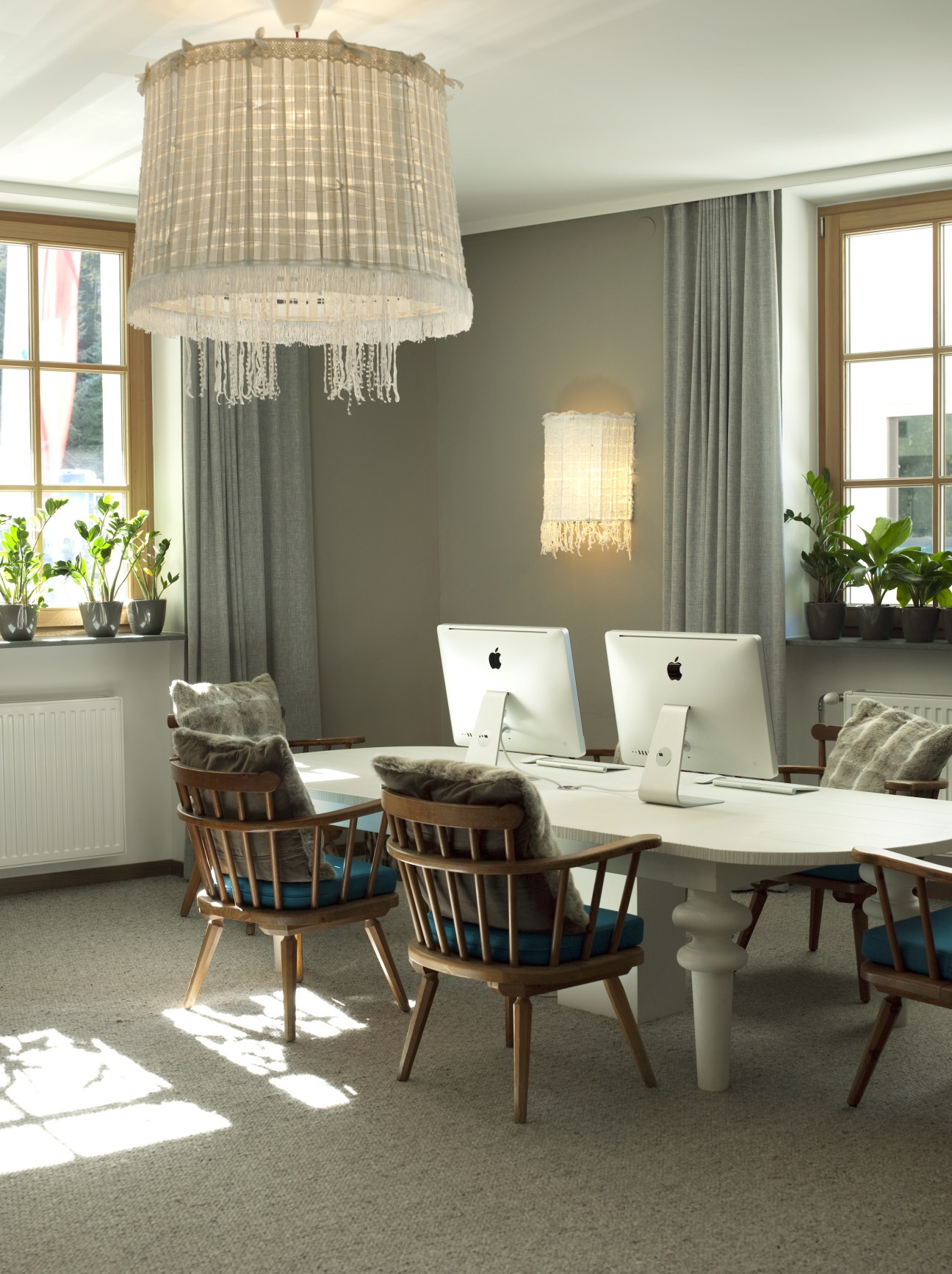 Modern hotel office space with wooden chairs, white desk, iMac computers, pendant lighting, and large windows.