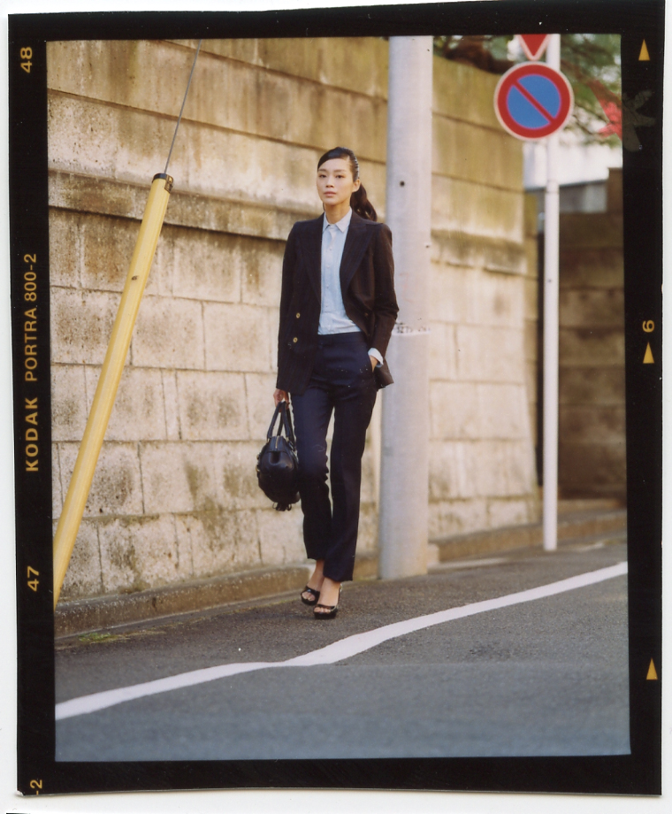 Woman in black blazer and trousers walking on Tokyo street past stone wall.