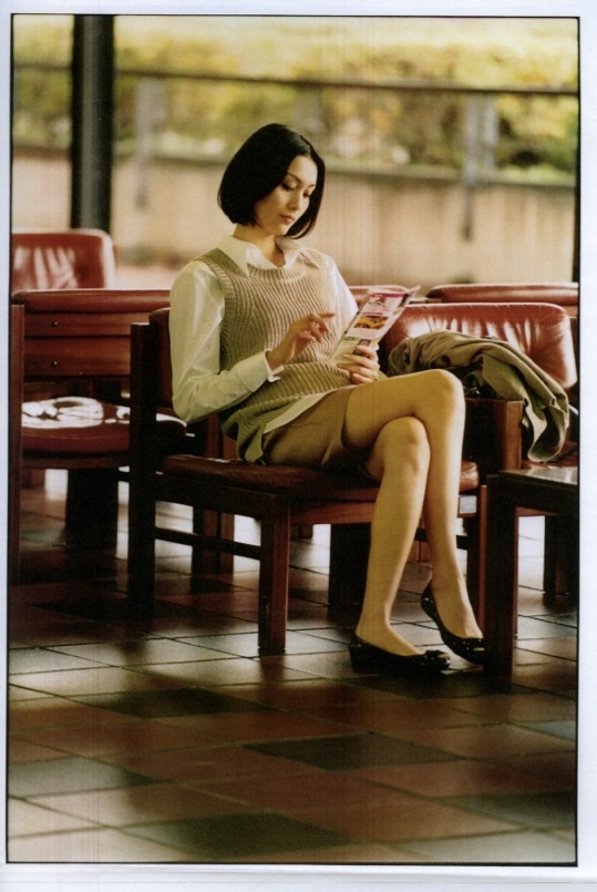 Woman in cream knit and olive shorts reading magazine on wooden bench indoors.