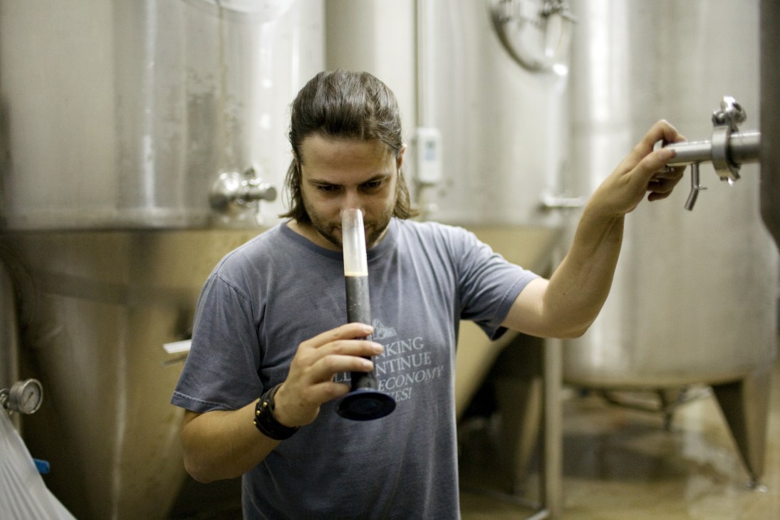 A brewery worker samples beer from a tap in an industrial facility with large metal tanks.