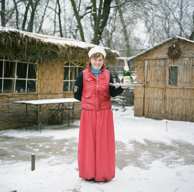 Smiling waitress in red vest and white headscarf holding a tray with wine bottle and glass outside snow-covered wooden restaurant structures.
