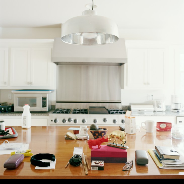 Objects arranged on a wooden kitchen table including fabric samples, brass animal figurines, books, and personal items beneath a modern pendant lamp.