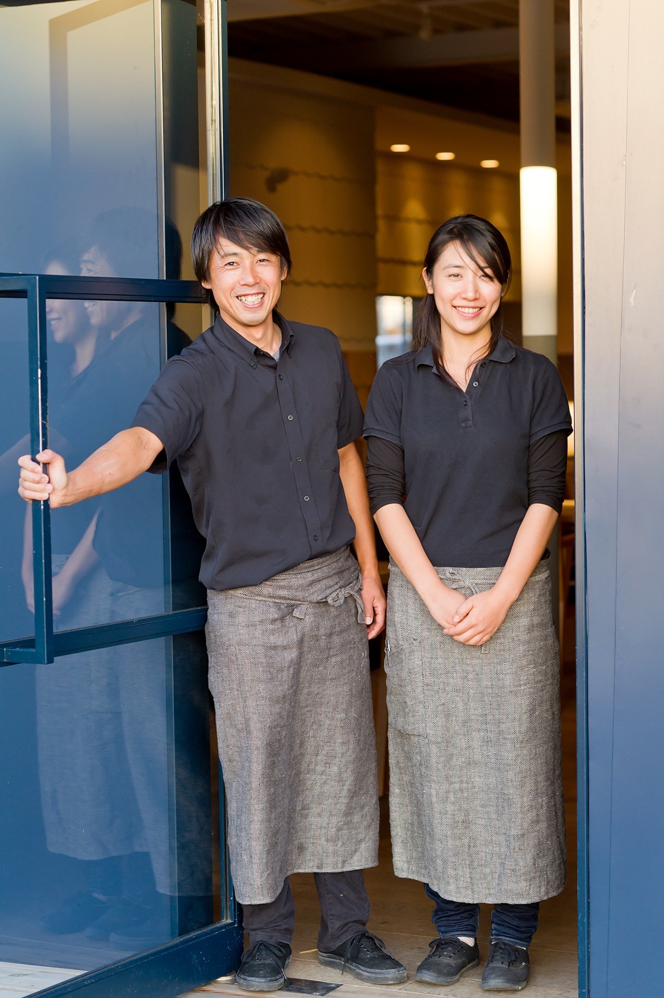 Two people in black shirts and gray aprons stand smiling in a doorway with blue frames.