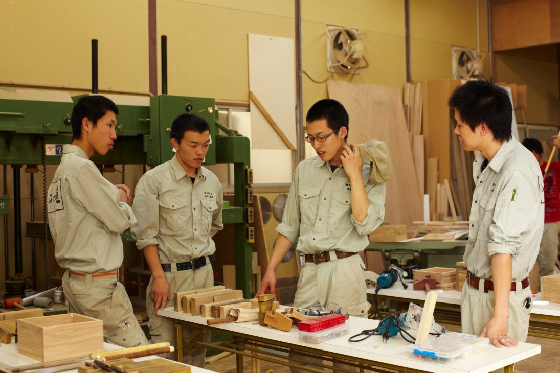 Four young apprentices in beige work shirts discuss woodworking in a workshop with machines and wooden materials.