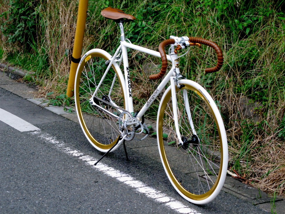 White Kinfolk Bicycles road bike with brown leather handlebars and gold wheels parked on a Tokyo street.