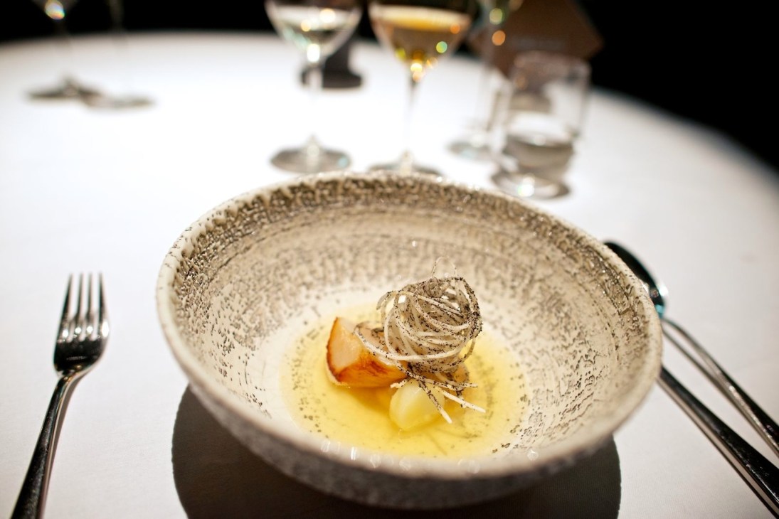 An elegantly plated fine dining dish in a textured ceramic bowl with sauce and garnish, place setting visible in background.