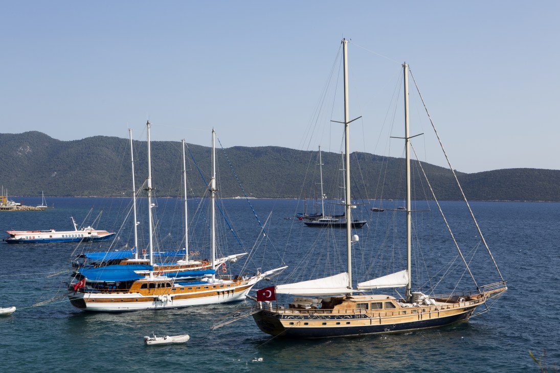 Multiple traditional wooden gulet sailing vessels anchored in turquoise water off Icmeler, Bodrum, with forested mountains in the background.