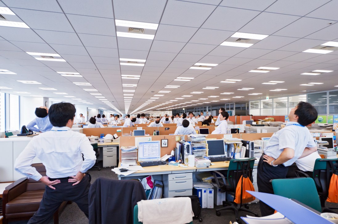 Office workers in white shirts performing synchronized stretches at their desks in a Mitsubishi Electric factory floor.