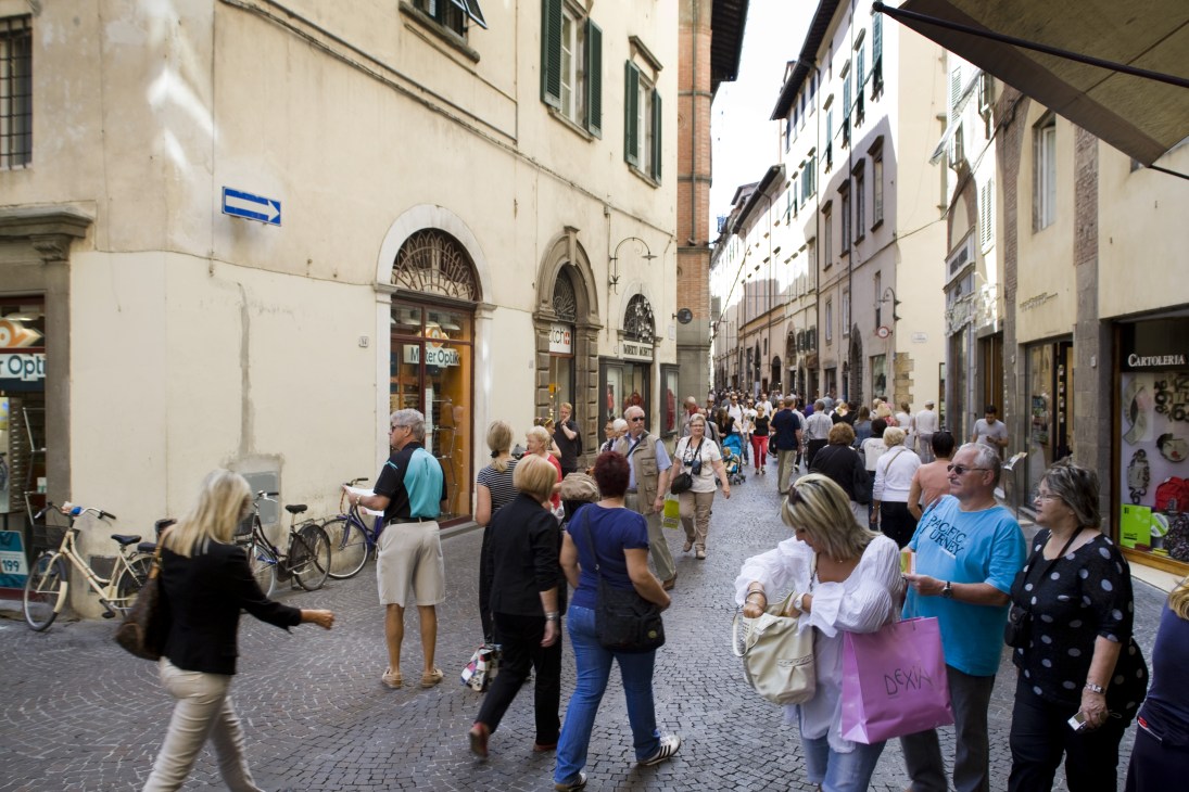 Pedestrians and cyclists on a busy cobblestone shopping street in Lucca lined with Renaissance buildings.