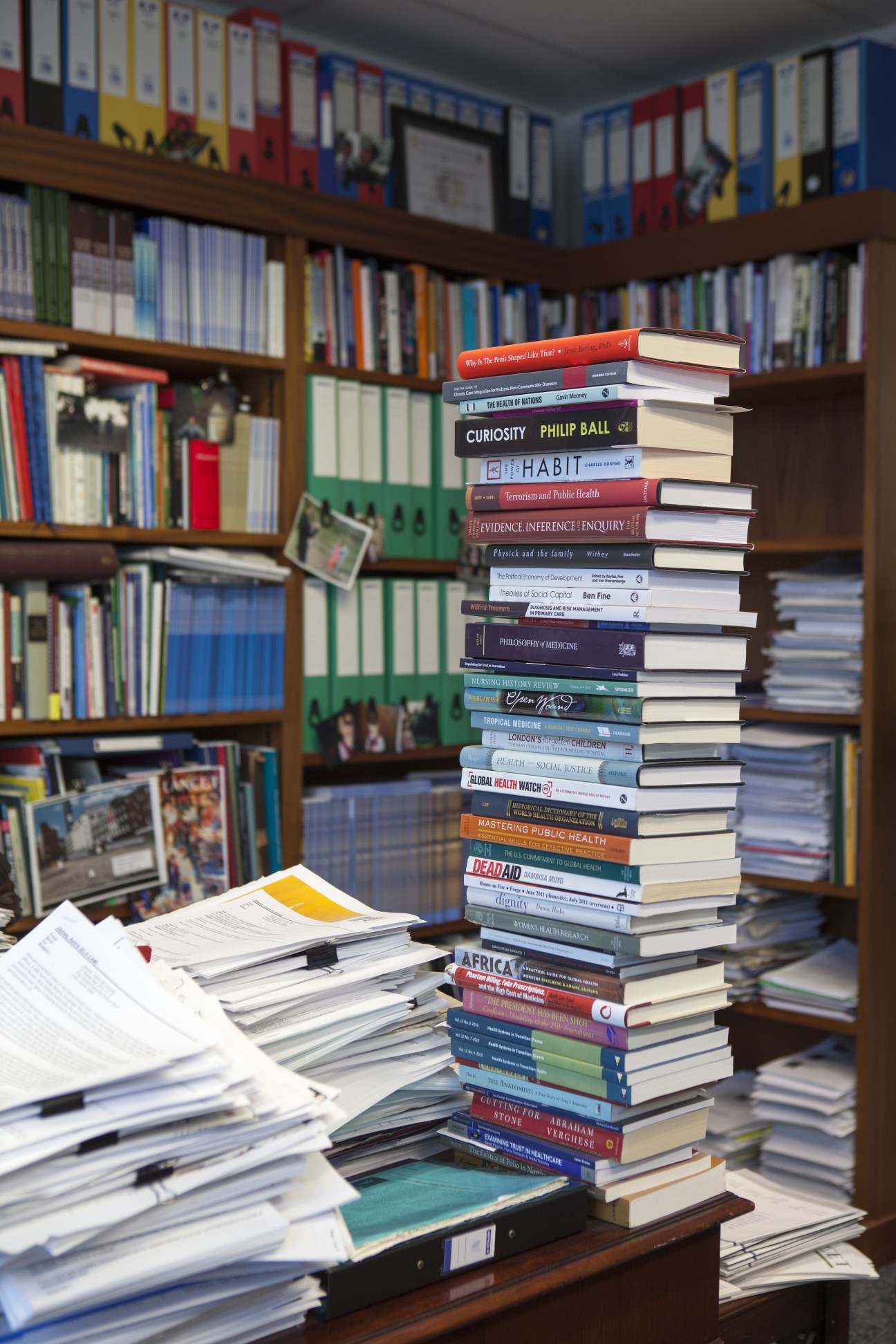 A tall, precarious stack of books piled on a desk in front of wooden bookshelves.
