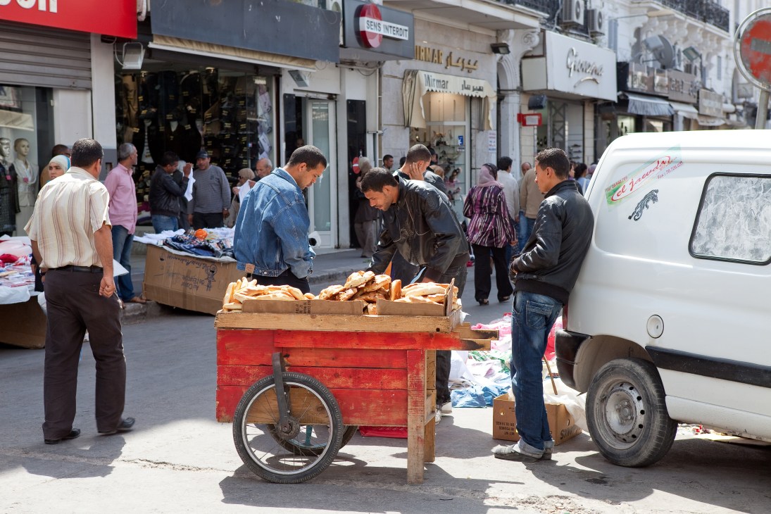 Street vendors sell bread from a red cart on a busy Tunis street lined with shops.