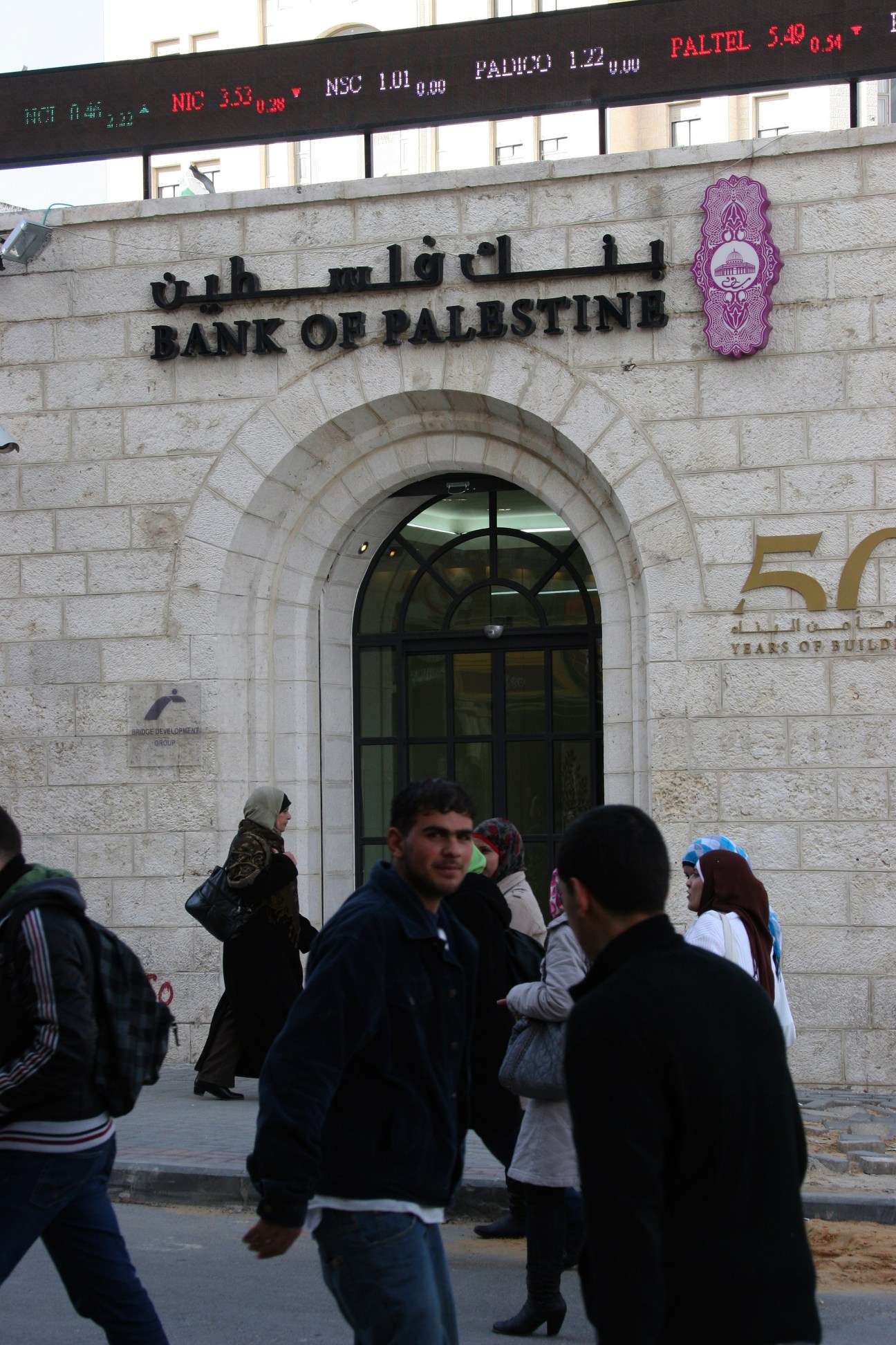 The Bank of Palestine storefront in Ramallah with Arabic and English signage, stock ticker above, and pedestrians outside.