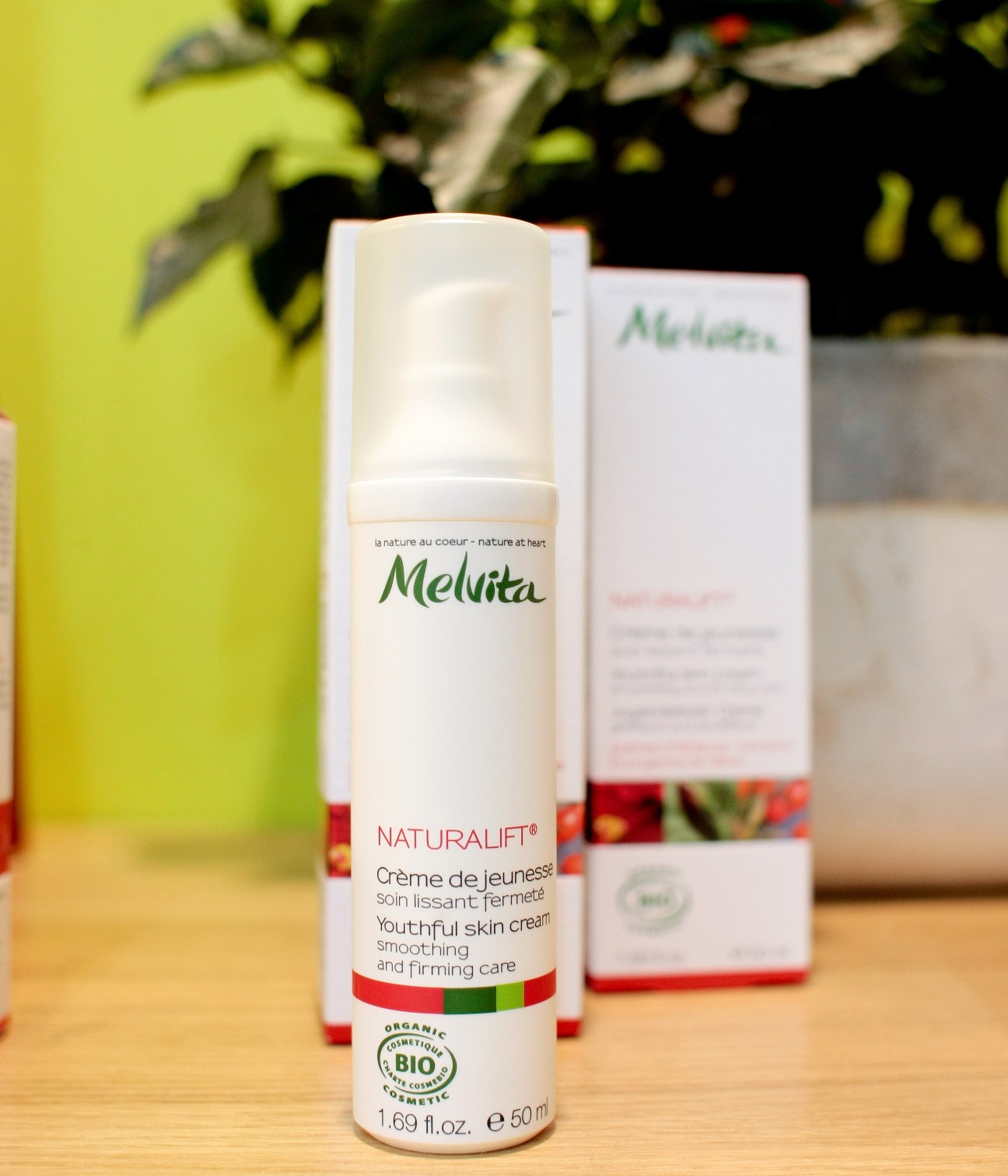 Melvita Naturalift organic youthful skin cream bottle with packaging displayed on wooden surface with plant in background.