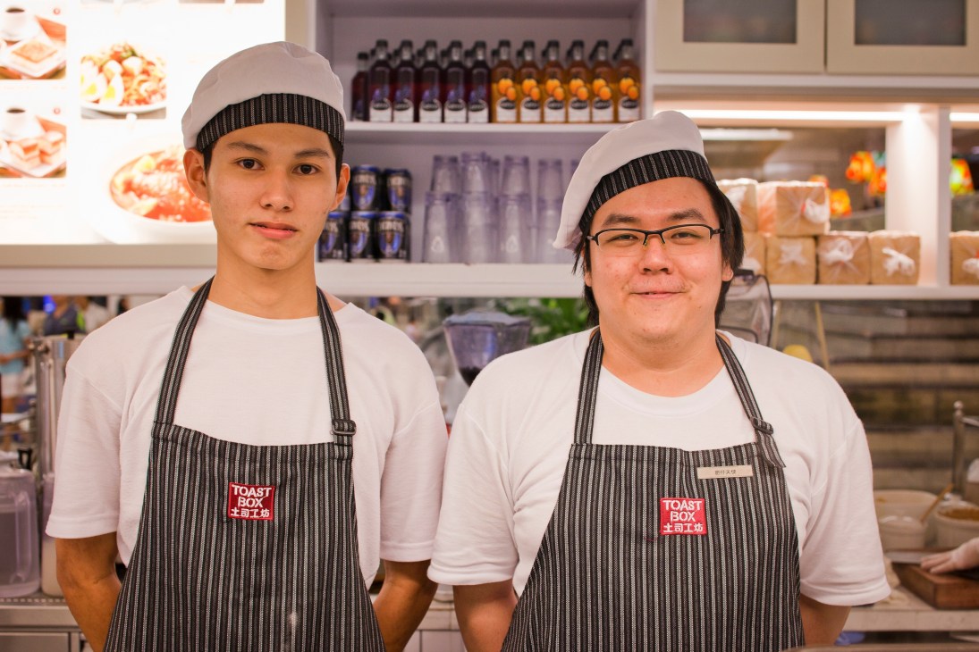 Two Toast Box staff members wearing striped aprons and caps stand in front of shelves of products.