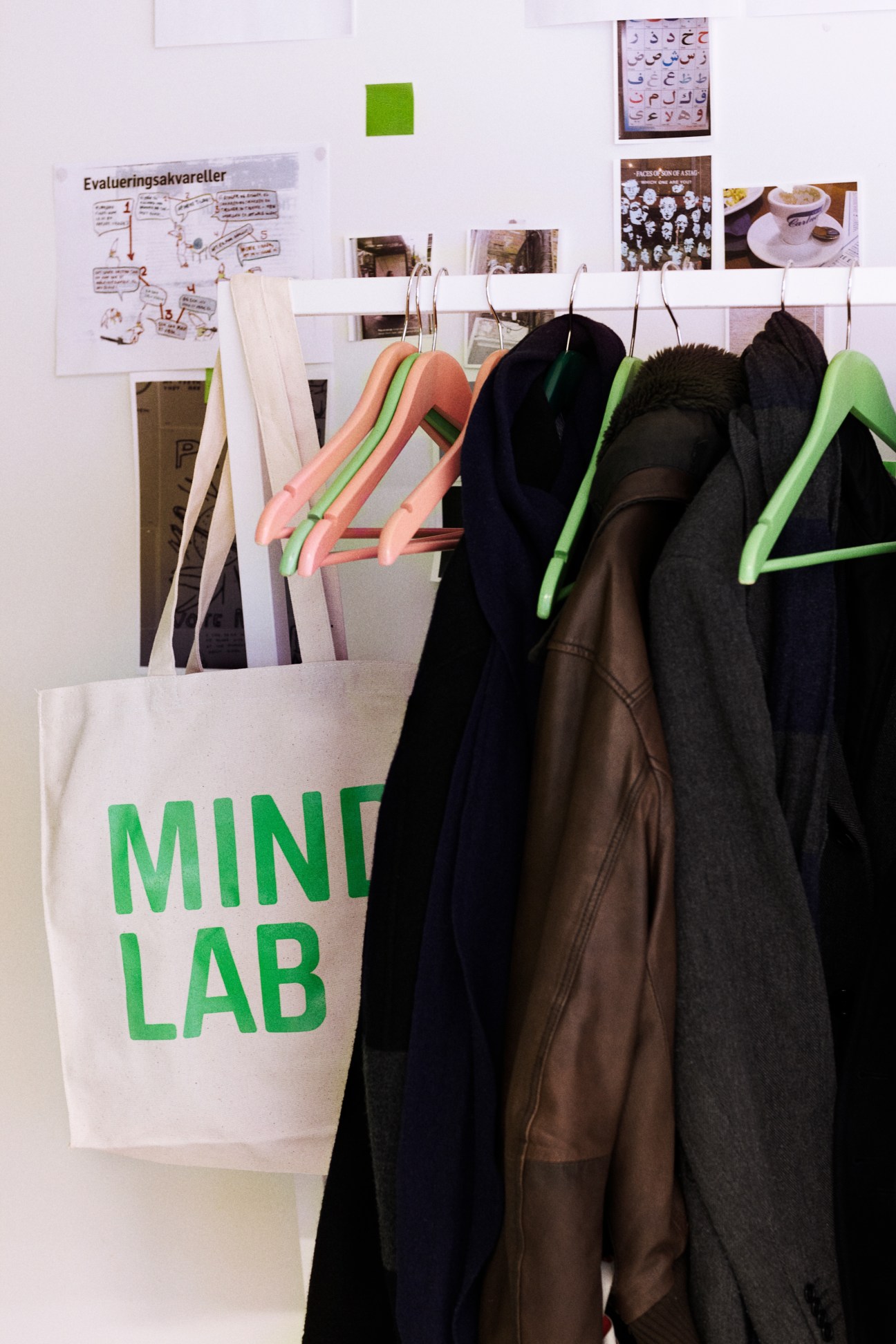 Mindlab office coatrack with colored hangers holding jackets next to a white tote bag and design sketches on wall.