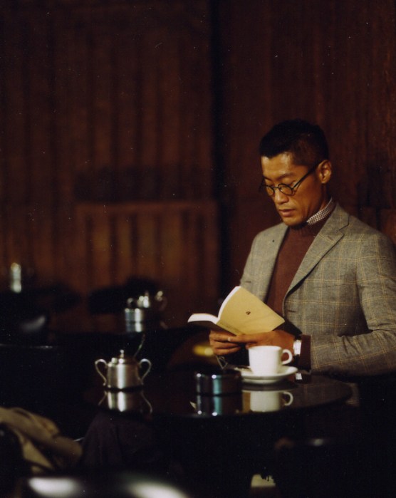 Man wearing Dunhill jacket and Oliver Peoples glasses reading at a café table.