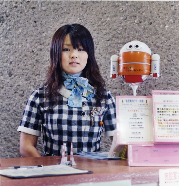 Young woman in blue plaid shirt with blue bow tie standing at space museum desk with robot mascot.