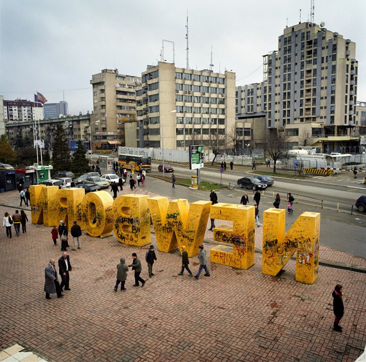 Yellow 'Newborn' monument spelling out large letters in Pristina's city square with Soviet-style apartment buildings and pedestrians in background.