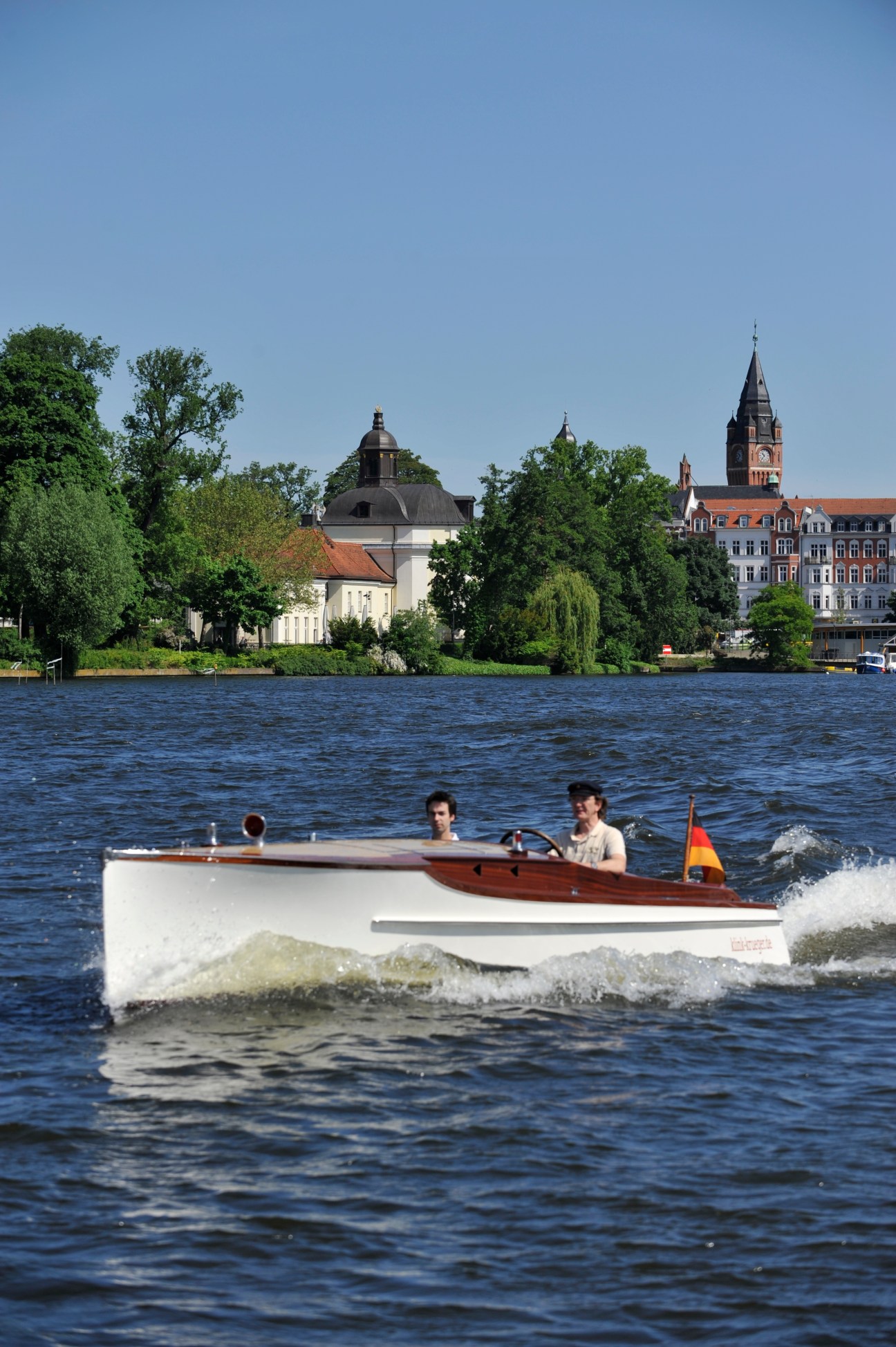 A wooden-hull motorboat with white sides and brown trim carrying three people on a lake in Berlin, with historic buildings in the background.
