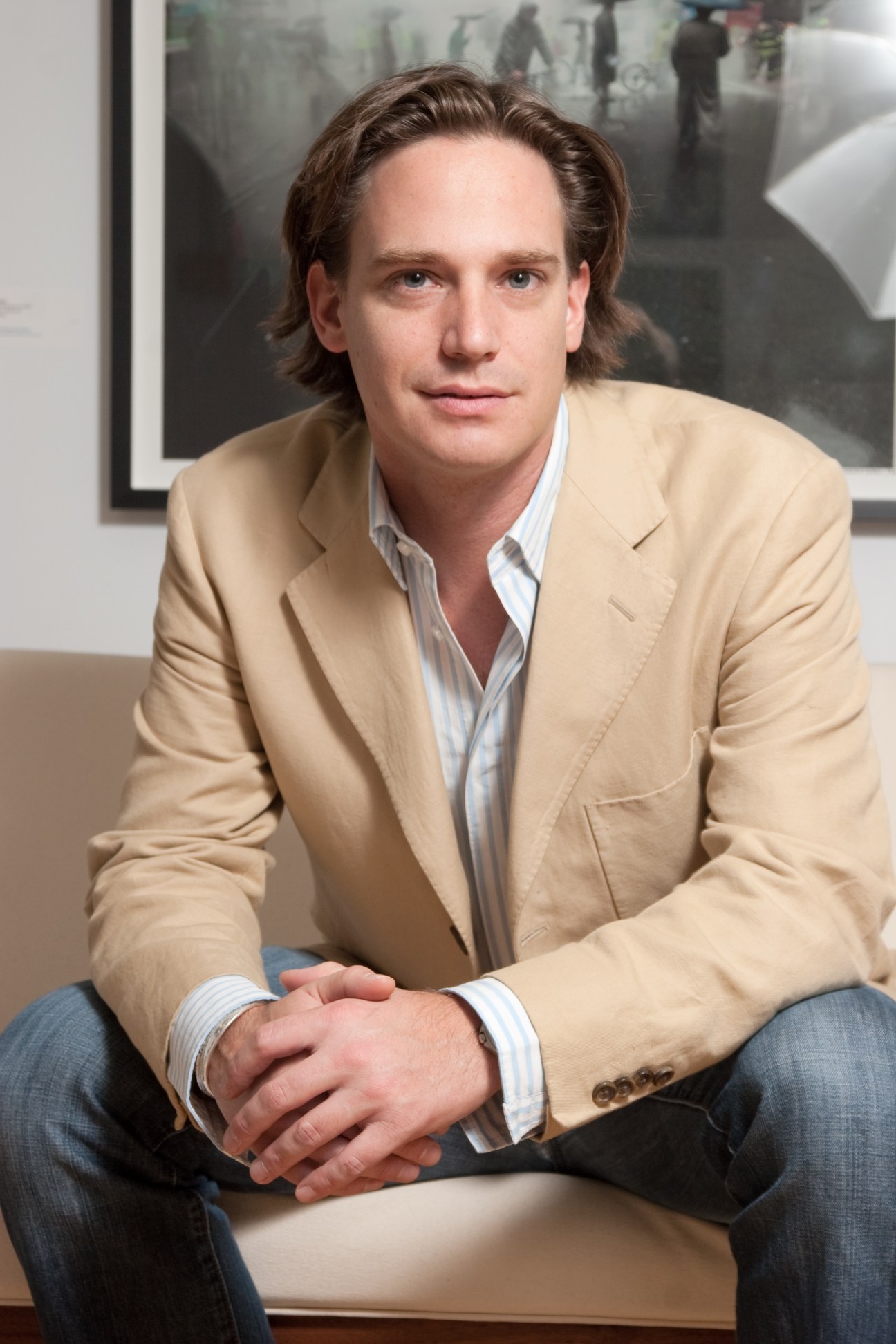 Man in beige blazer and striped shirt sitting on beige sofa with framed artwork visible behind him