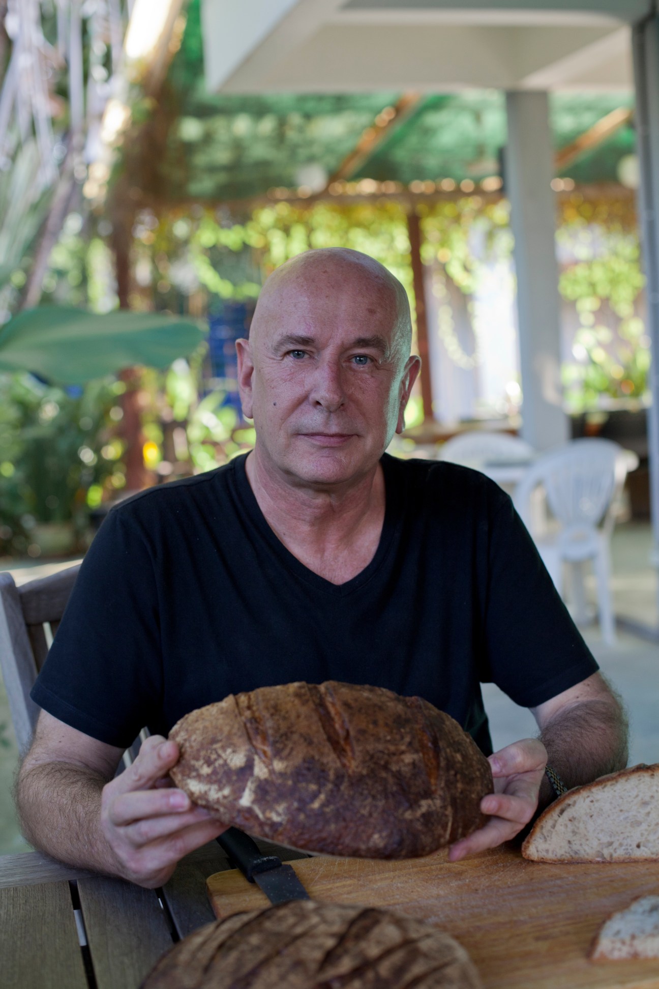 Michael Conkey, a baker in Bangkok, holds a round loaf of sourdough bread at a table with greenery in the background.