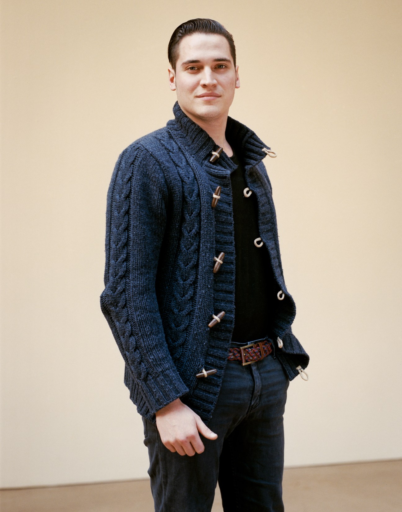 Thomas Lazzarini, BA Product Design student from Italy, wears a navy cable-knit cardigan over a black shirt against a beige background.