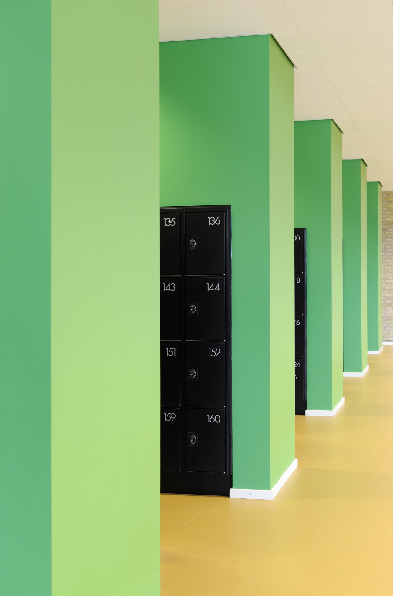 Black numbered lockers mounted on a bright green wall in a school corridor with natural wood flooring.