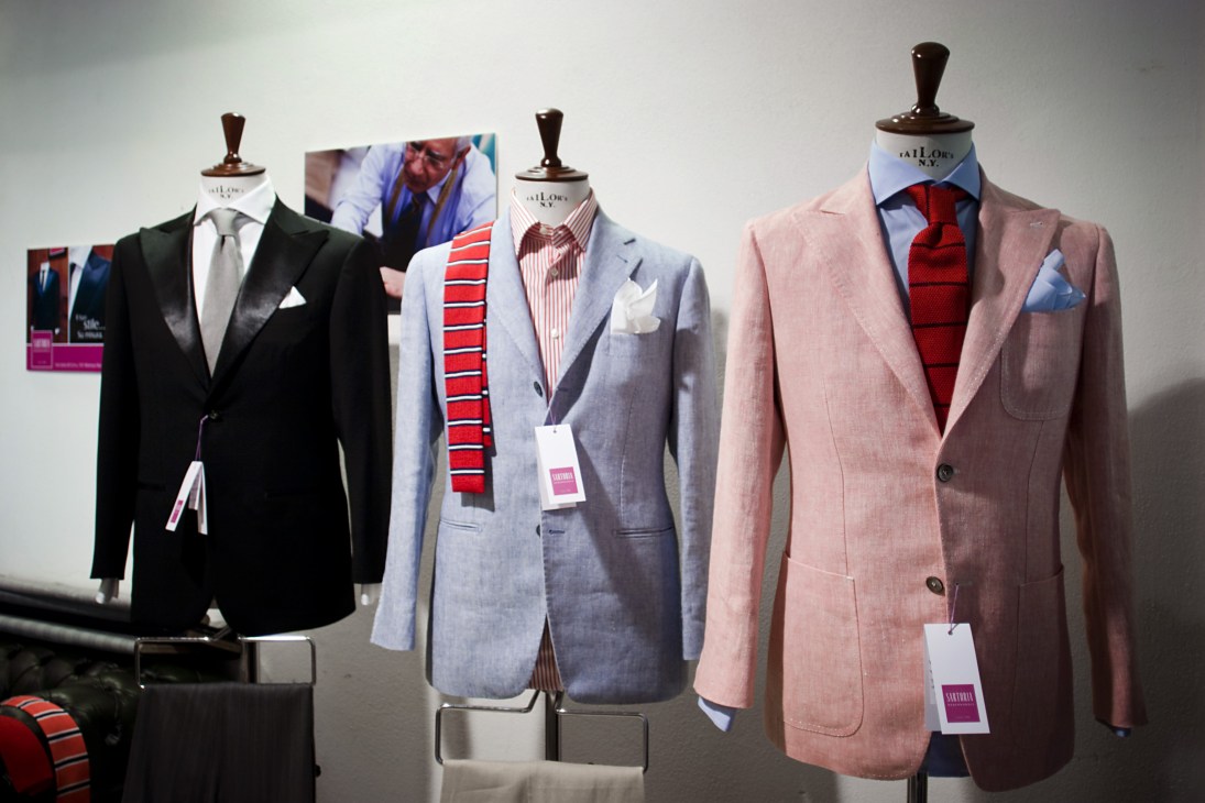 Three tailored blazers on dress forms display unlined jackets in black, light blue, and pink with coordinating shirts and ties.
