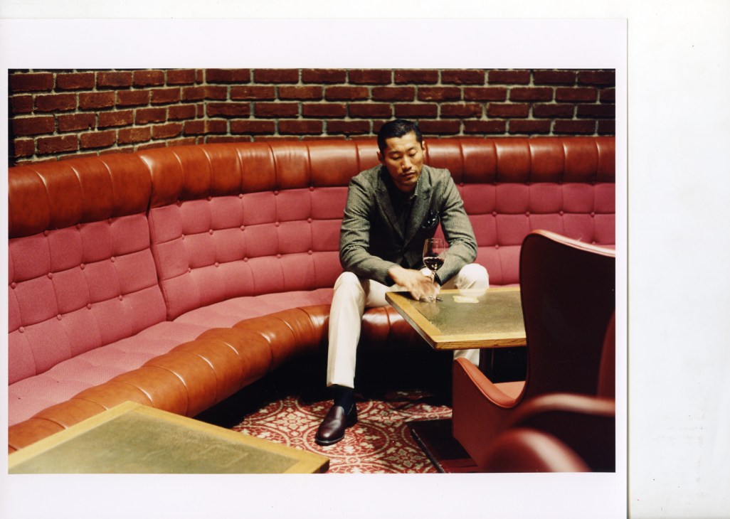Man seated on red leather banquette in retro 1970s bar setting with brick wall, holding a drink.