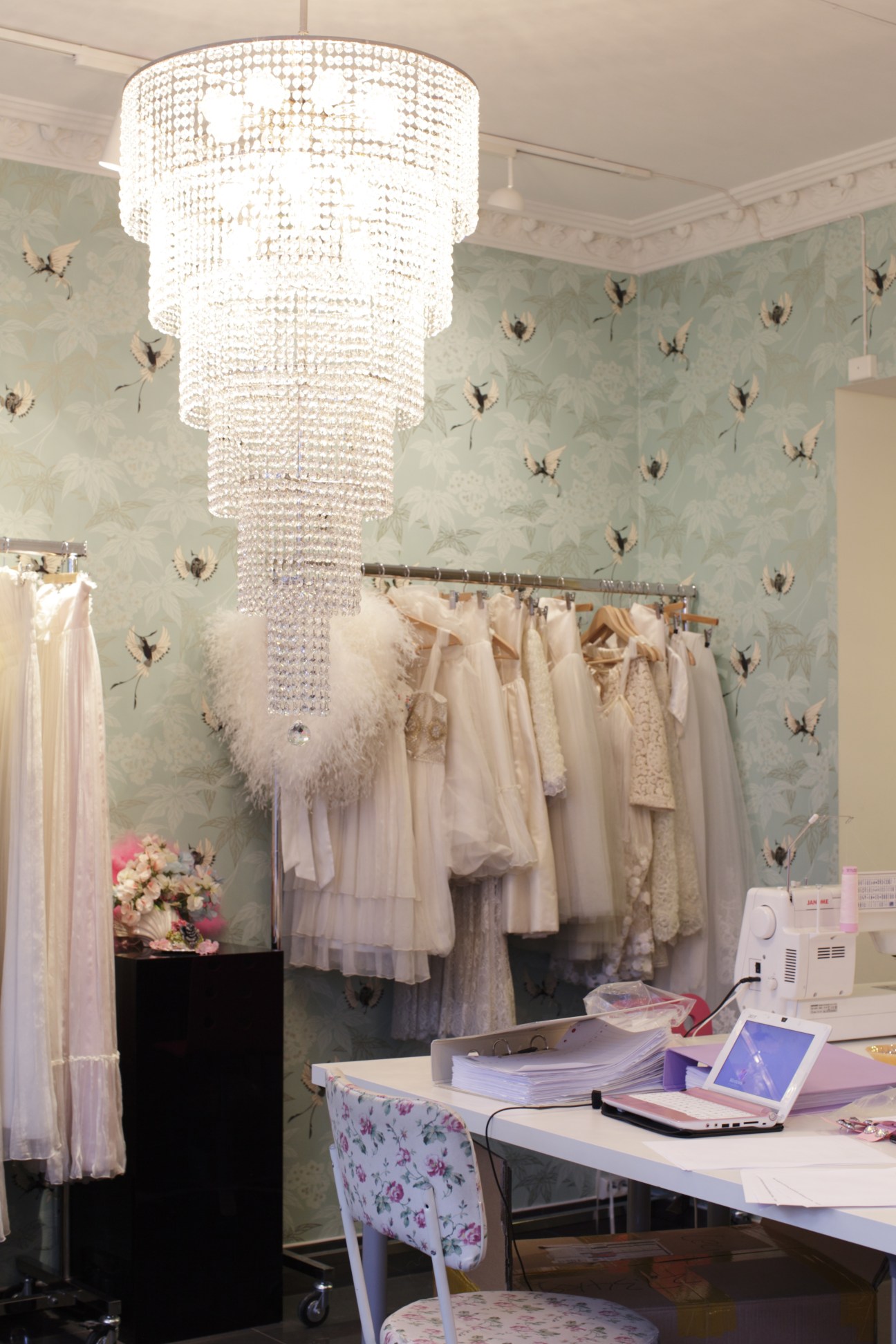 Interior of Cecilie Melli's fashion studio and shop featuring a crystal chandelier, pale dresses on a rack, and wallpaper with botanical motifs.
