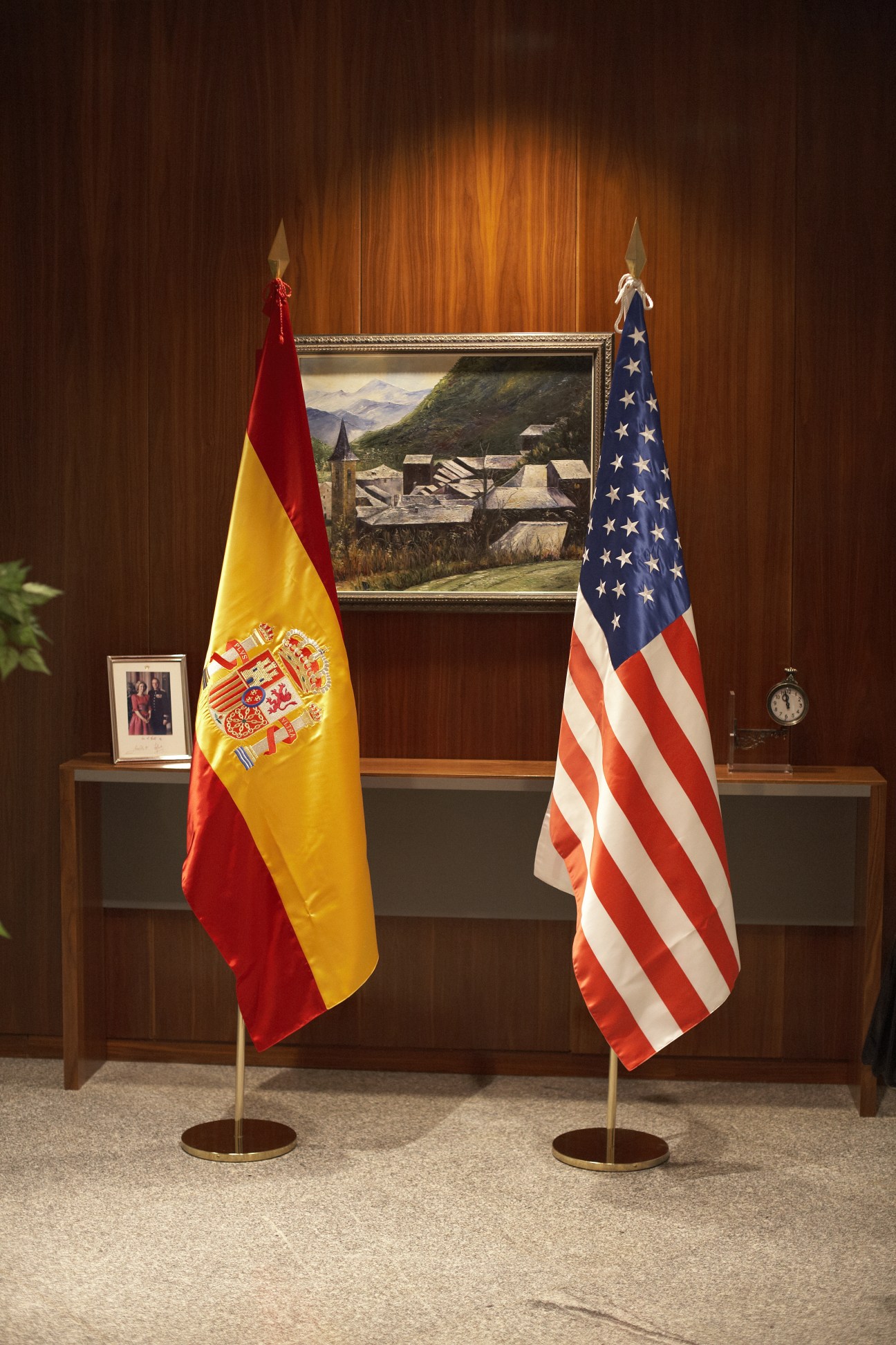 Spanish and American flags flanking a framed landscape painting in a formal wood-paneled room.