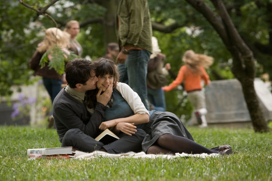 A young couple sits on grass in a park, reading together while others enjoy the outdoor setting in the background.