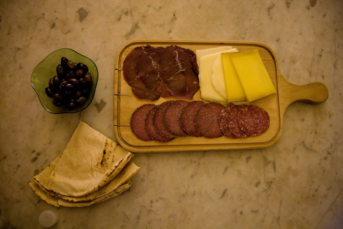 Black olives in a green bowl, sliced cured meats and cheese on a wooden board, with bread on a marble surface.