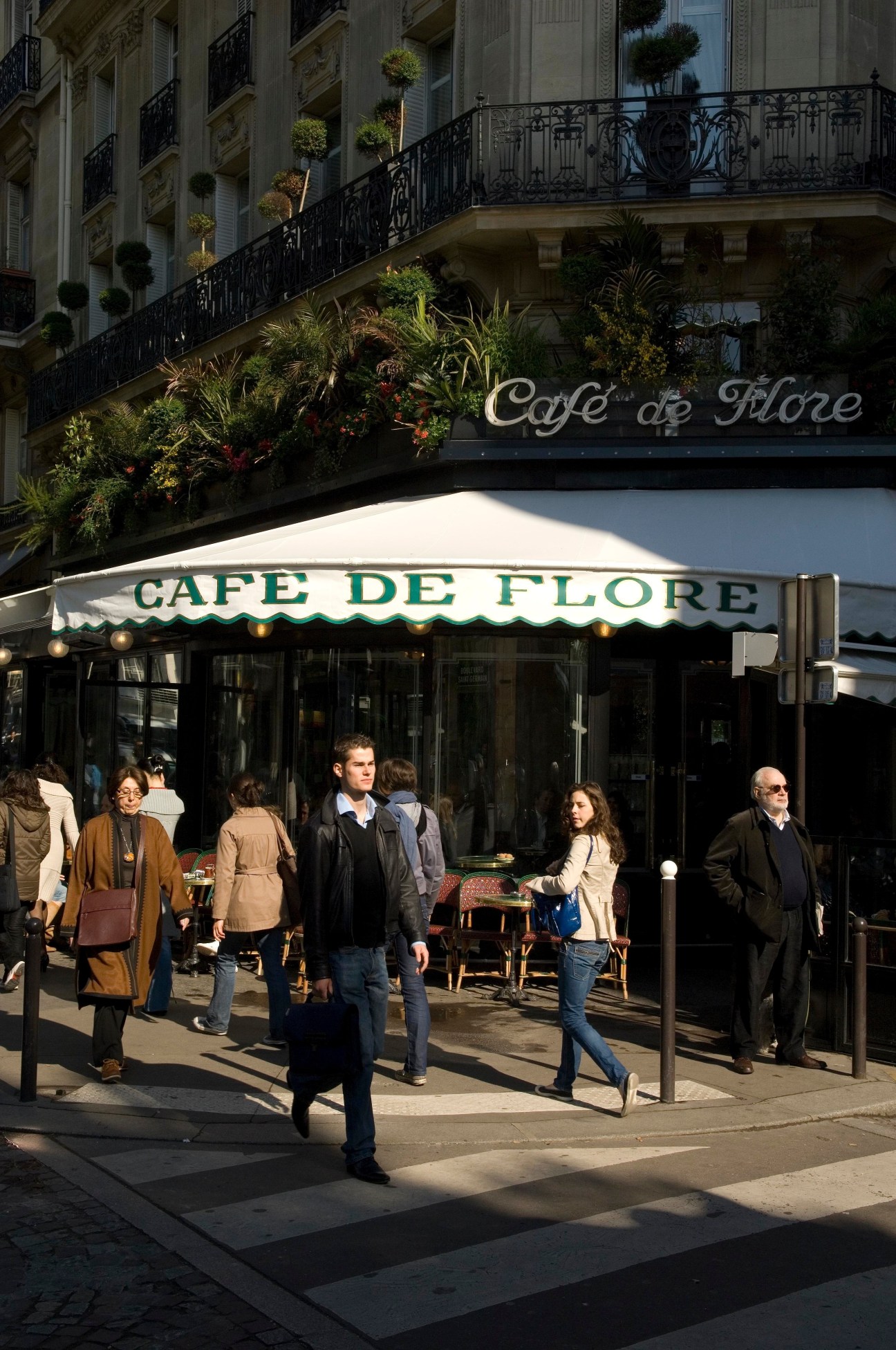 Café de Flore's corner storefront with green and white awning, ornate Parisian building facade, and pedestrians on the street.