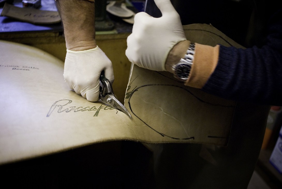 Hands in white gloves cutting leather shoe sole material with scissors.
