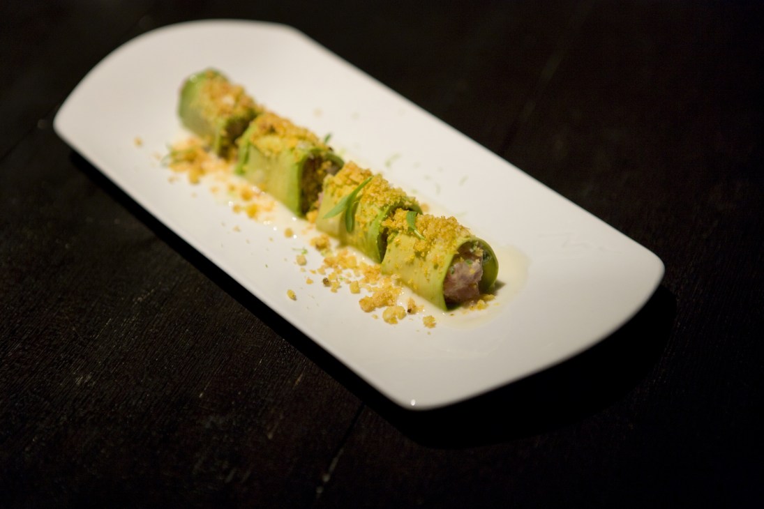 Ceviche and avocado sushi rolls garnished with micro cilantro and gold crumble on a white rectangular plate.