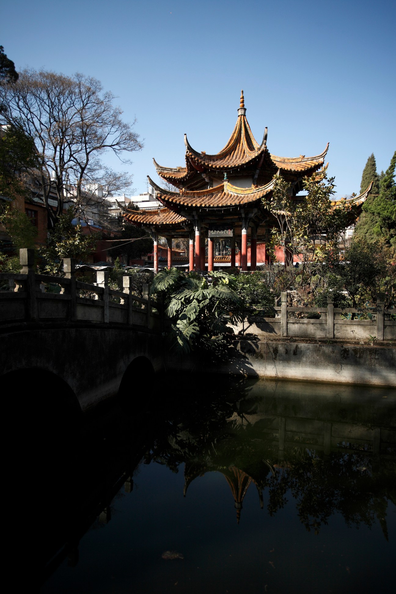 Traditional Chinese pagoda with golden tiered roofs reflected in still water beside a bridge and garden.