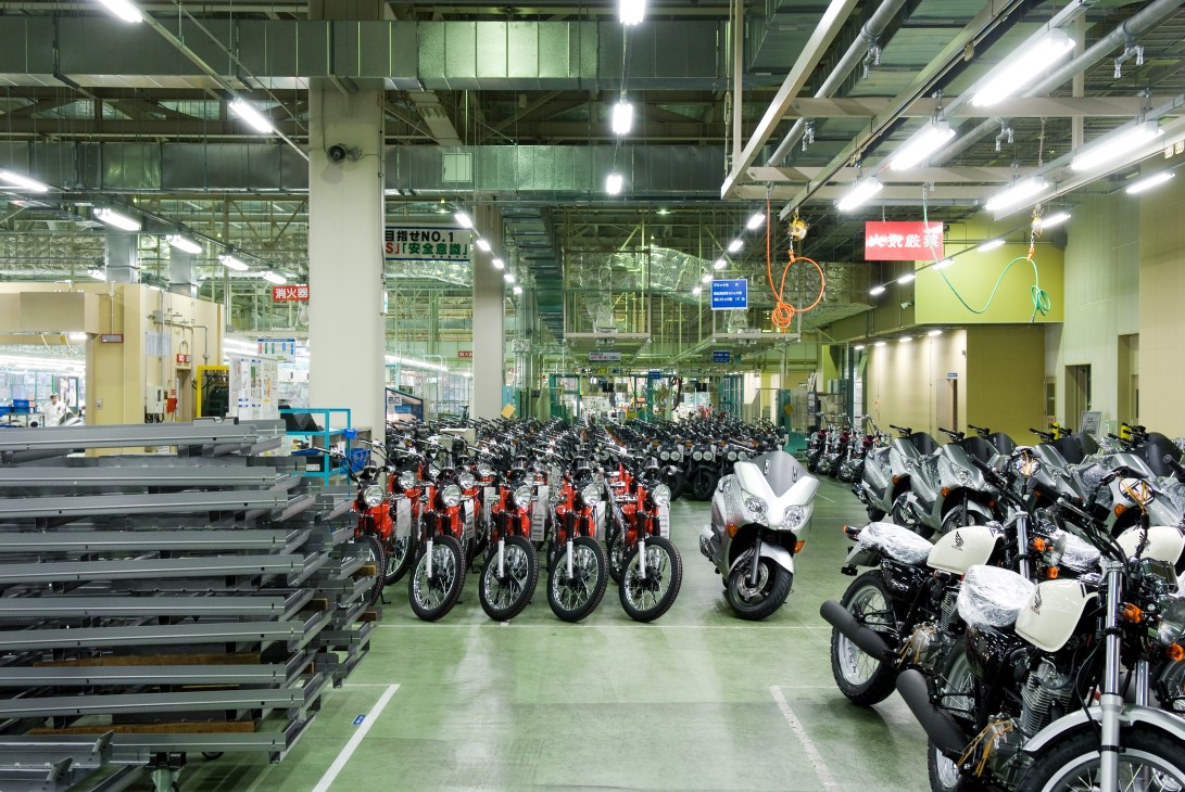 Rows of finished red and white Honda scooters lined up in a factory warehouse with metal framework and industrial lighting.