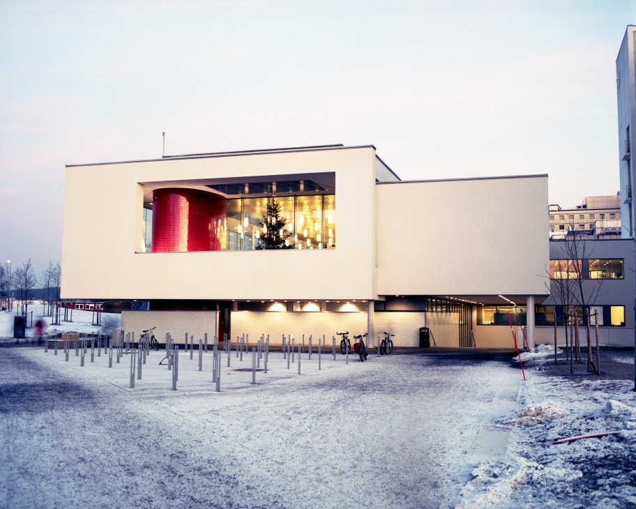 Modern hospital building exterior with cream-colored facade, large illuminated windows, and red cylindrical architectural element visible inside.