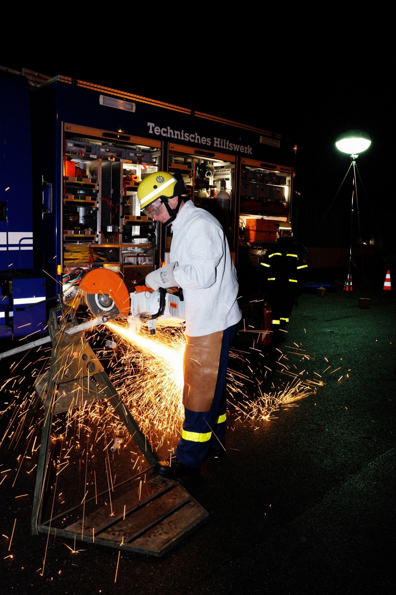 THW volunteer in yellow hard hat and safety gear operating a circular saw at night next to equipment truck.