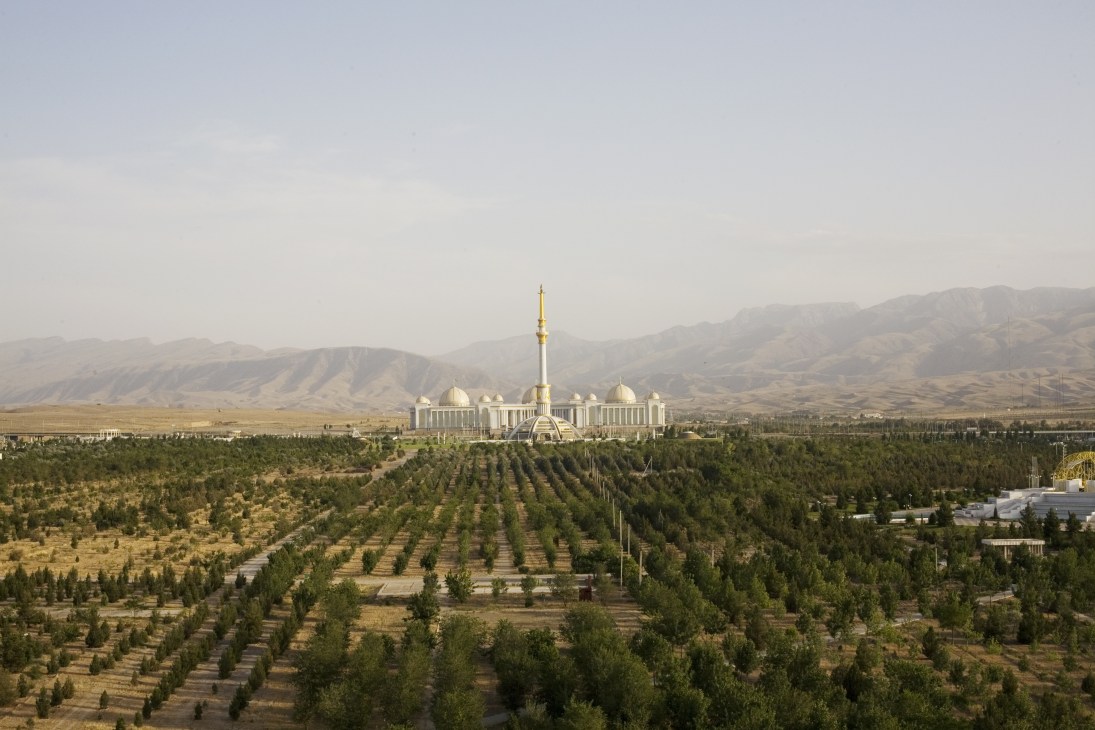White mosque with golden dome and minaret surrounded by planted trees and mountains in Ashgabat, Turkmenistan.